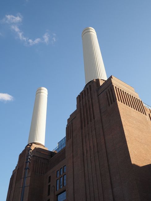 A large historic power station building constructed from red brick, prominently featuring four tall, beige smokestacks extending upward from its roof. The building is situated along the edge of a rive