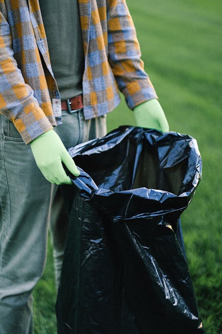 A man with short dark hair, wearing a black T-shirt and light blue jeans, stands next to a large wheeled rubbish cart filled with flattened cardboard boxes, some partially wrapped in plastic. He is ou