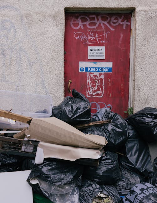 A small, light blue pickup truck parked on a street with its flatbed and cargo area overflowing with various types of waste and rubbish items. The truck’s cargo includes cardboard boxes, black plast